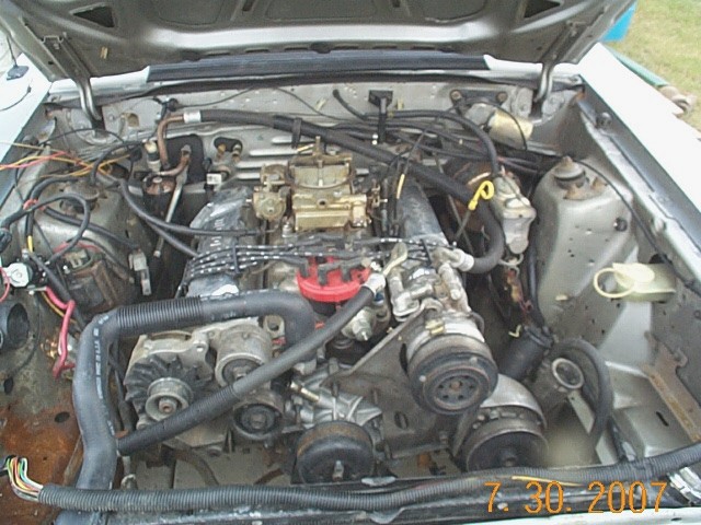 Engine bay of silver 1984 Ford Mustang with carbureted V8 and red distributor cap