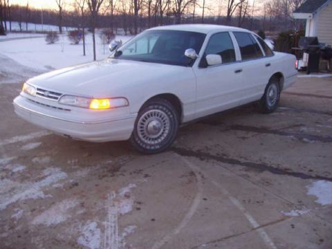1996 Ford Crown Victoria - A white sedan is parked on a dirt lot with snow on the ground and trees in the background.