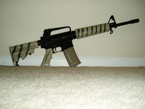 1995 Mercury Grand Marquis - An assault rifle with a tiger stripe camouflage pattern applied to its stock and barrel shroud rests on a carpeted floor.