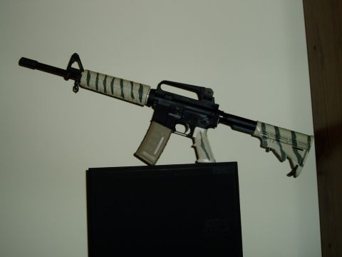 1995 Mercury Grand Marquis - An assault rifle with tiger stripe camouflage tape on its barrel and stock is shown against a light-colored wall.