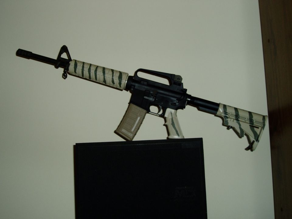 Assault rifle with tiger stripe tape on barrel and stock, tan magazine, and white grip, displayed on a black surface