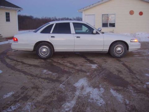 1996 Ford Crown Victoria - A white 1996 Ford Crown Victoria sedan is parked on a dirt lot next to a house.