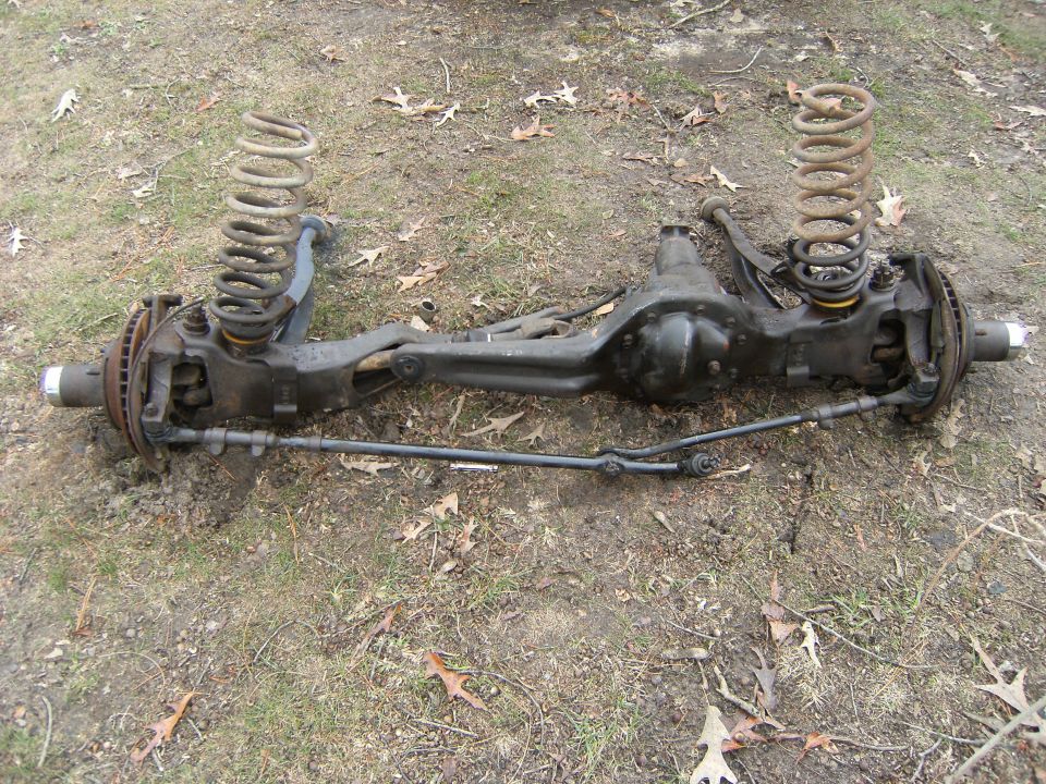Removed front solid drive axle with coil springs and tie rod lying on grass