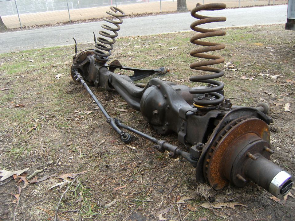 Removed solid front axle with coil springs, tie rod and rusty brake rotor on grass