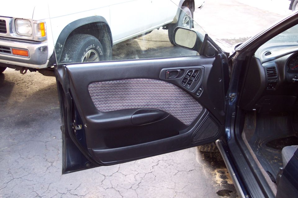 Interior view of a dark-colored car door panel with patterned fabric insert and power window controls. A white pickup truck is visible outside