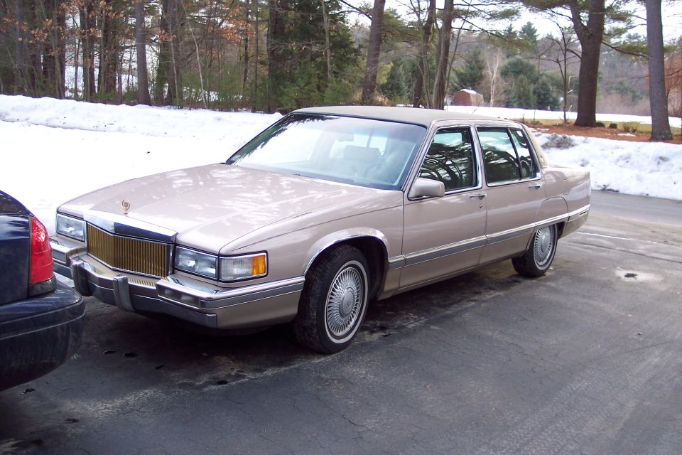 Tan Cadillac sedan parked next to a dark-colored car. Snow is visible in the background