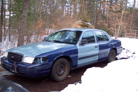 2001 Ford Crown Victoria - A two-tone Ford Crown Victoria sits in the snow, with one headlight missing and the front bumper showing signs of damage.