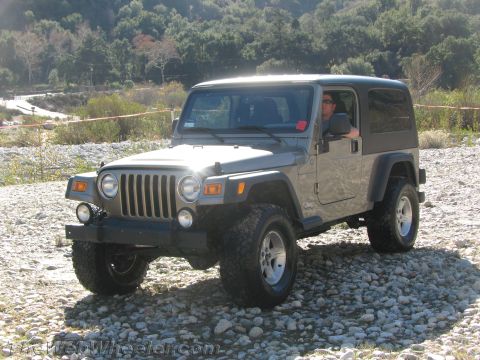 1987 Suzuki Samurai - A driver sits in a gray Jeep Wrangler parked on a gravelly terrain with a wooded hillside behind it.