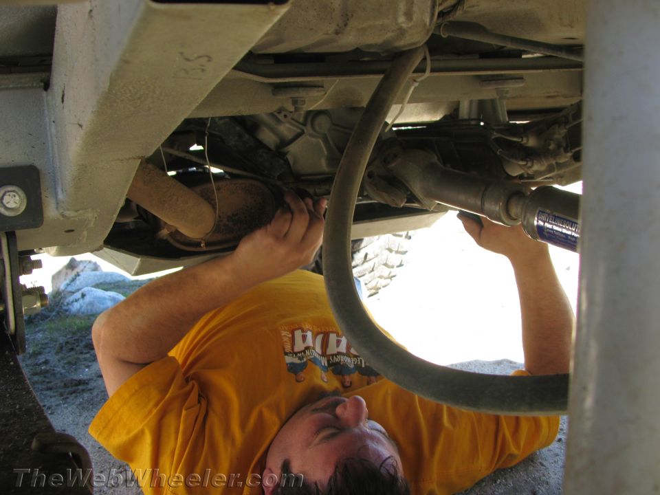 Underside of a vehicle with a person in a yellow shirt working on the drivetrain and exhaust system