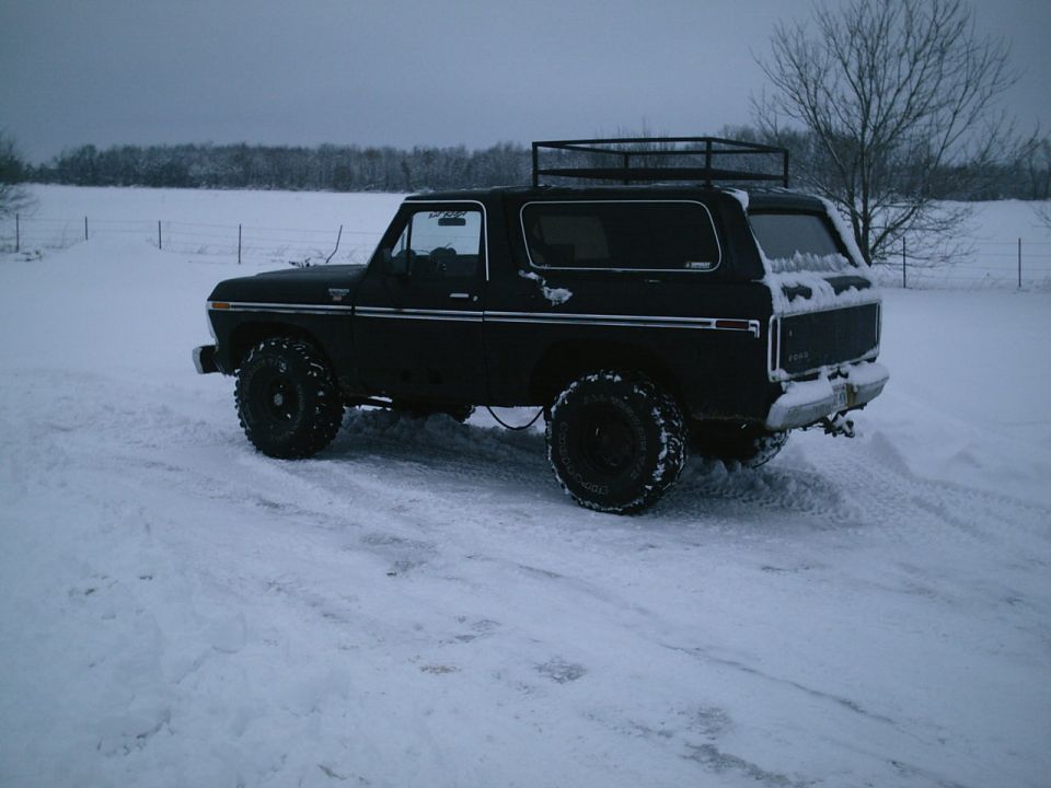Black lifted Ford Bronco with roof rack and big off-road tires parked in snow, rear three-quarter view
