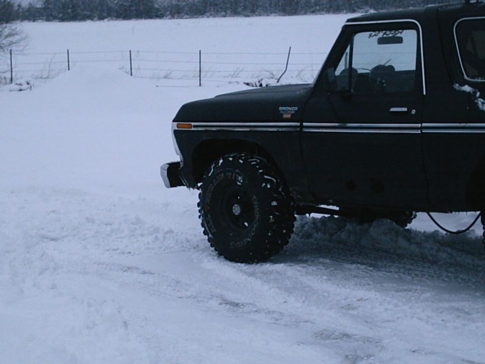 Dark Ford Bronco with lift and large all-terrain tires parked in snow, side view of front half