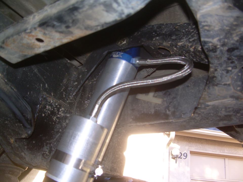 Close-up of a silver shock absorber with a blue top and braided hose, mounted to vehicle undercarriage