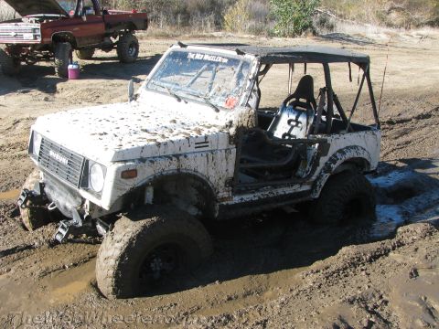 1987 Suzuki Samurai - A white 1987 Suzuki Samurai is stuck deep in mud, with another red truck visible in the background.
