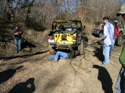 1996 Ford Bronco - A group of people are gathered around a yellow Jeep Wrangler on a dirt path, with one person working underneath it from underneath.