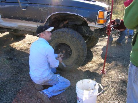 1996 Ford Bronco - A man works on the front wheel of a muddy Ford Bronco II, with another person operating a jack.