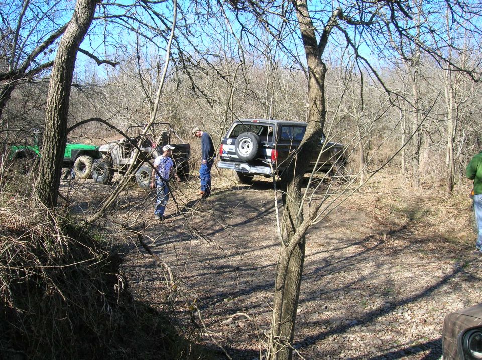 Several off-road vehicles parked on a dirt path in a wooded area with people standing around