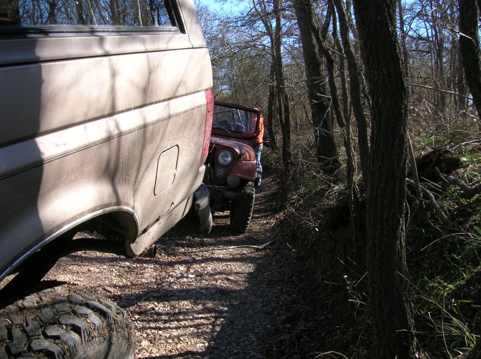 Tan Ford Bronco rear quarter panel and red Jeep front side on a dirt path in woods