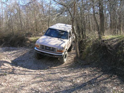 1996 Ford Bronco - A tan 1996 Ford Bronco navigates a wooded area with a gravelly, uneven terrain.