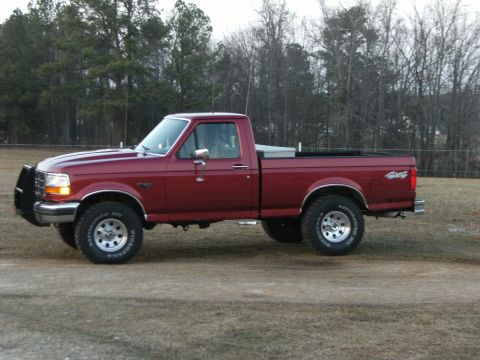 1992 Ford F-150 - Full driver-side profile of a dark red Ford F-150 regular-cab long-bed 4x4 parked on dry winter grass in front of a pine treeline. Visible…