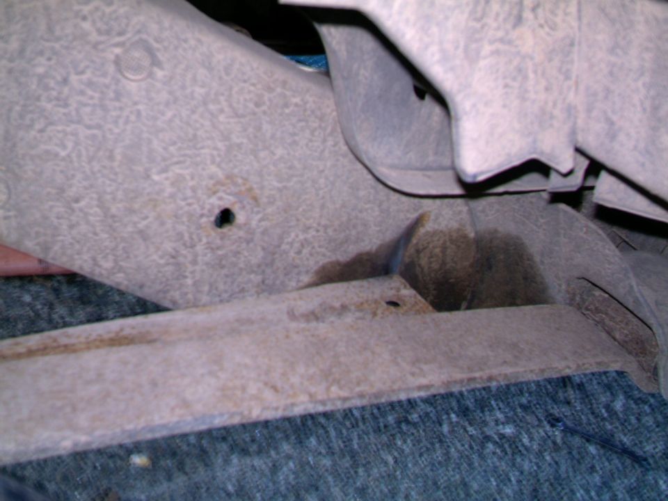 Bare rusty sheet metal floor bracing with drilled hole above gray carpet