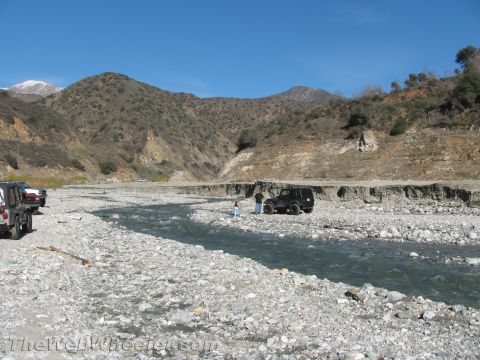 1987 Suzuki Samurai - Two vehicles are visible in a dry riverbed with mountains under a clear blue sky.