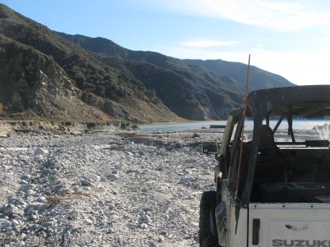 1987 Suzuki Samurai - A white 1987 Suzuki Samurai is parked on a rocky shore, with mountains and a lake visible in the distance.