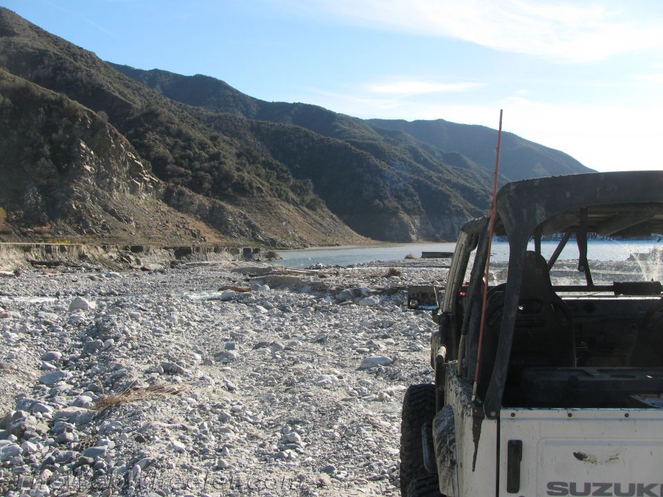White 1987 Suzuki Samurai rear view parked on rocky shore with mountains and lake in background