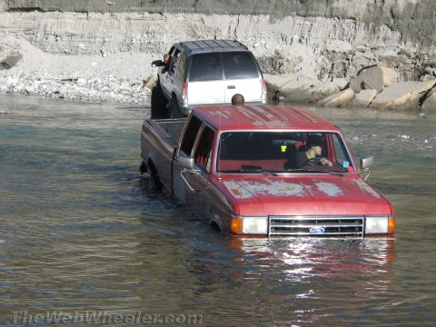 1987 Suzuki Samurai - Two vehicles navigate a shallow river, with the red pickup truck in the foreground partially submerged.