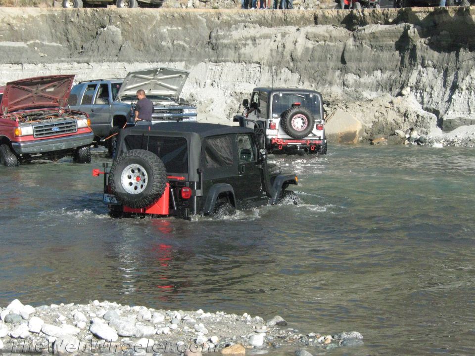 Several Jeeps and a red Ford pickup truck are driving through a river with rocky banks