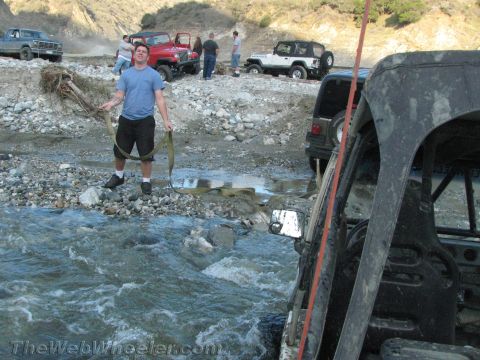 1987 Suzuki Samurai - Several off-road vehicles are gathered in a rocky stream bed, with a man holding a tow strap in the foreground.