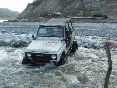 1987 Suzuki Samurai - A white 1987 Suzuki Samurai navigates a rocky riverbed, with other vehicles visible on the bank in the distance.