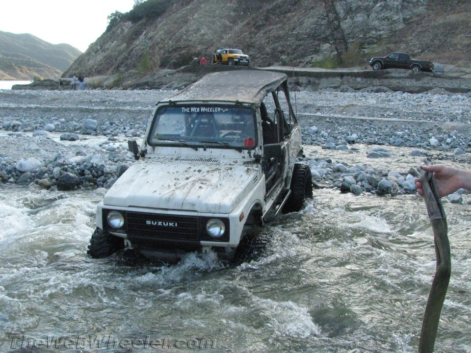 White 1987 Suzuki Samurai driving through a rocky river, with other vehicles in the background