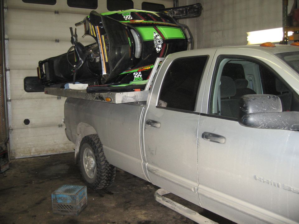 Silver Dodge Ram pickup with green snowmobile loaded on bed rack in a garage