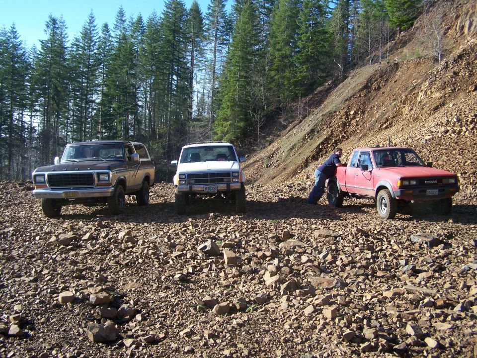 1978 Ford Bronco, white pickup, and red Nissan pickup on a rocky slope with trees
