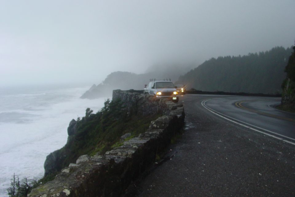 White SUV parked on a stone wall next to a winding road. Another vehicle is visible behind it. Ocean waves crash below