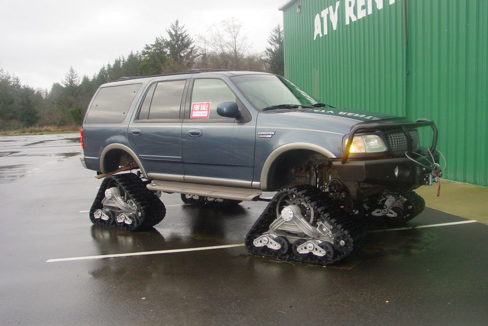 Blue SUV with tracks instead of wheels, parked in lot. Has a 'For Sale' sign in window