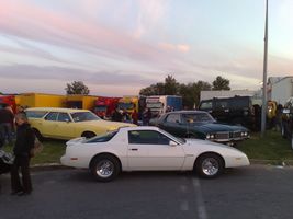 1991 Ford Crown Victoria - A white sports car is parked at an outdoor event alongside other vehicles and trailers under a colorful sky.