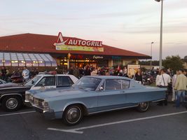 1991 Ford Crown Victoria - A light blue 1966 Chrysler Newport is parked in front of an Autogrill, with other vehicles and people visible.