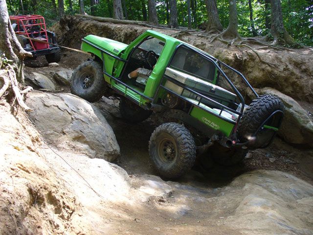 Green off-road truck climbing over rocks, with another red off-road vehicle behind it being pulled by a rope