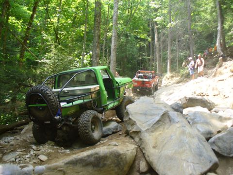1978 Ford Bronco - Two off-road vehicles tackle a challenging rocky trail in a wooded area.