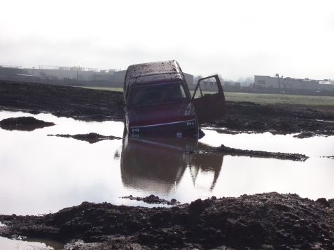 1986 Ford Bronco - A maroon van is partially submerged in a muddy puddle with its driver's side door open.