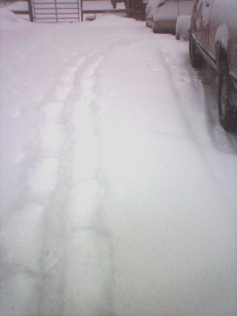 Snowy driveway with tire tracks and a red vehicle partially visible on the right