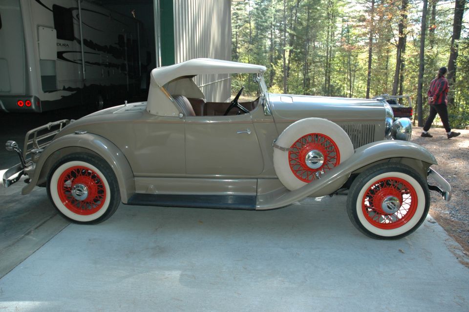Tan 1930s roadster in side view with red wire wheels and whitewall tires, parked near woods.