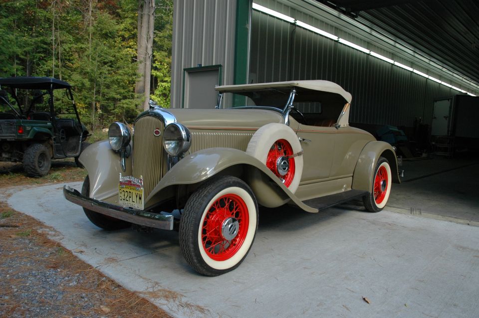Tan 1930s roadster with red wire wheels and whitewall tires parked by a metal shed