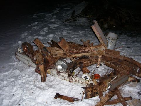 1979 Ford Pickup - A heavily rusted vehicle frame, possibly from the 1970s, lies in the snow with its headlights visible.