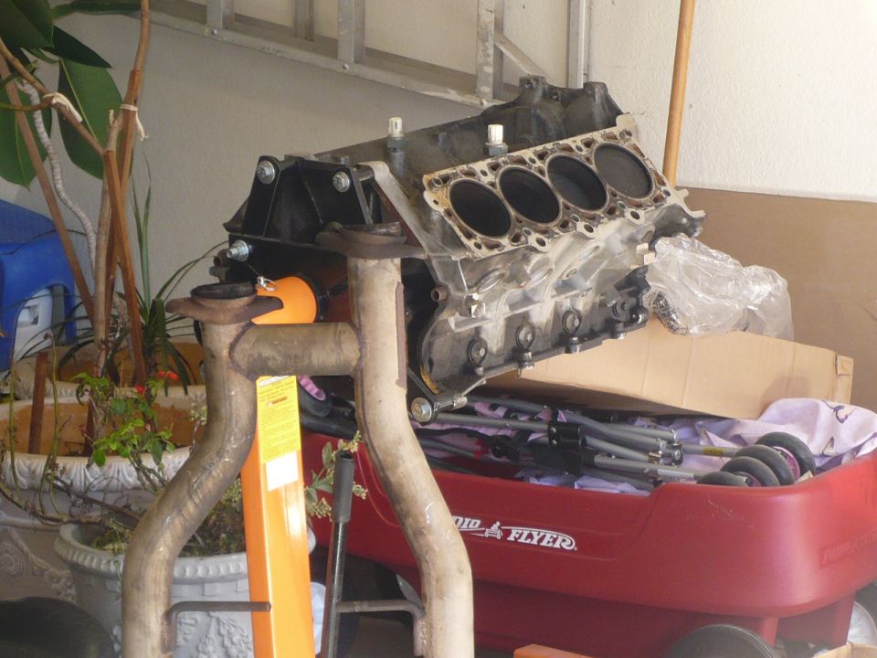 Engine block and exhaust pipes on an engine stand, with a red wagon full of parts in the background