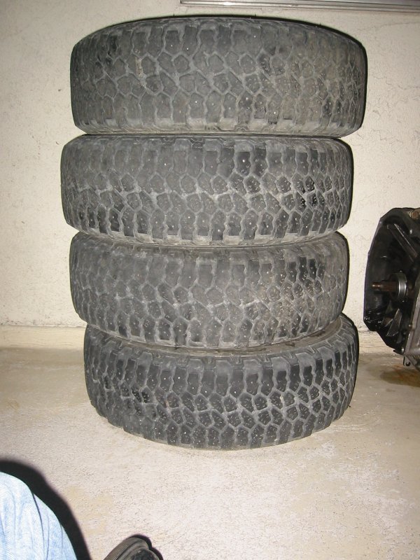 Stack of four worn all-terrain tires against a garage wall beside a transmission case