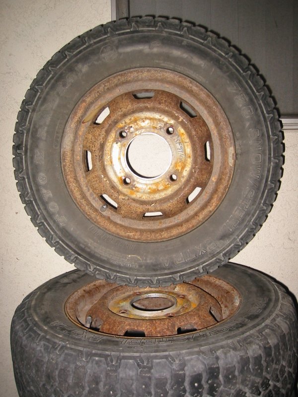 Two worn Toyo snow tires on rusty slotted steel wheels stacked against a wall