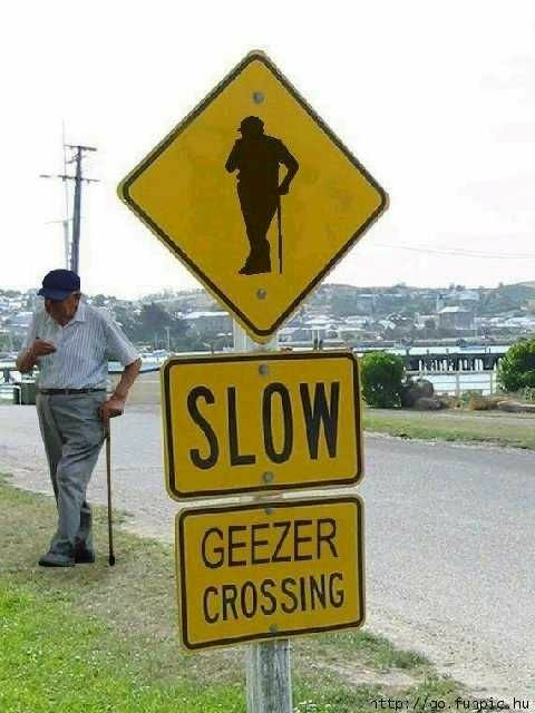 1972 Ford Ranchero - A man pauses near a "SLOW GEEZER CROSSING" sign, with a distant town visible.