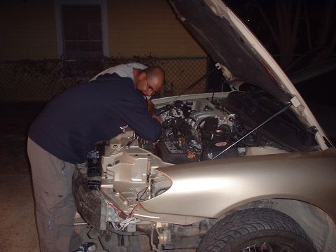 2000 Ford Taurus - A man works on the engine of a gold 2000 Ford Taurus, with the front fender removed and wires visible.
